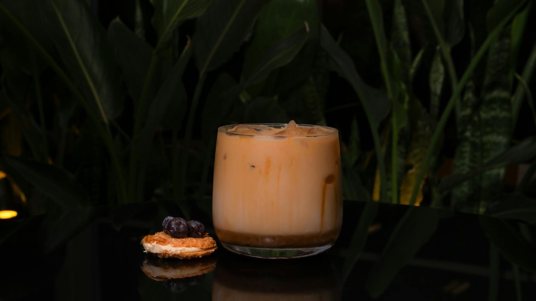 Mrożona kawa z ciastkiem i owocami na czarnym blacie wśród zielonych roślin