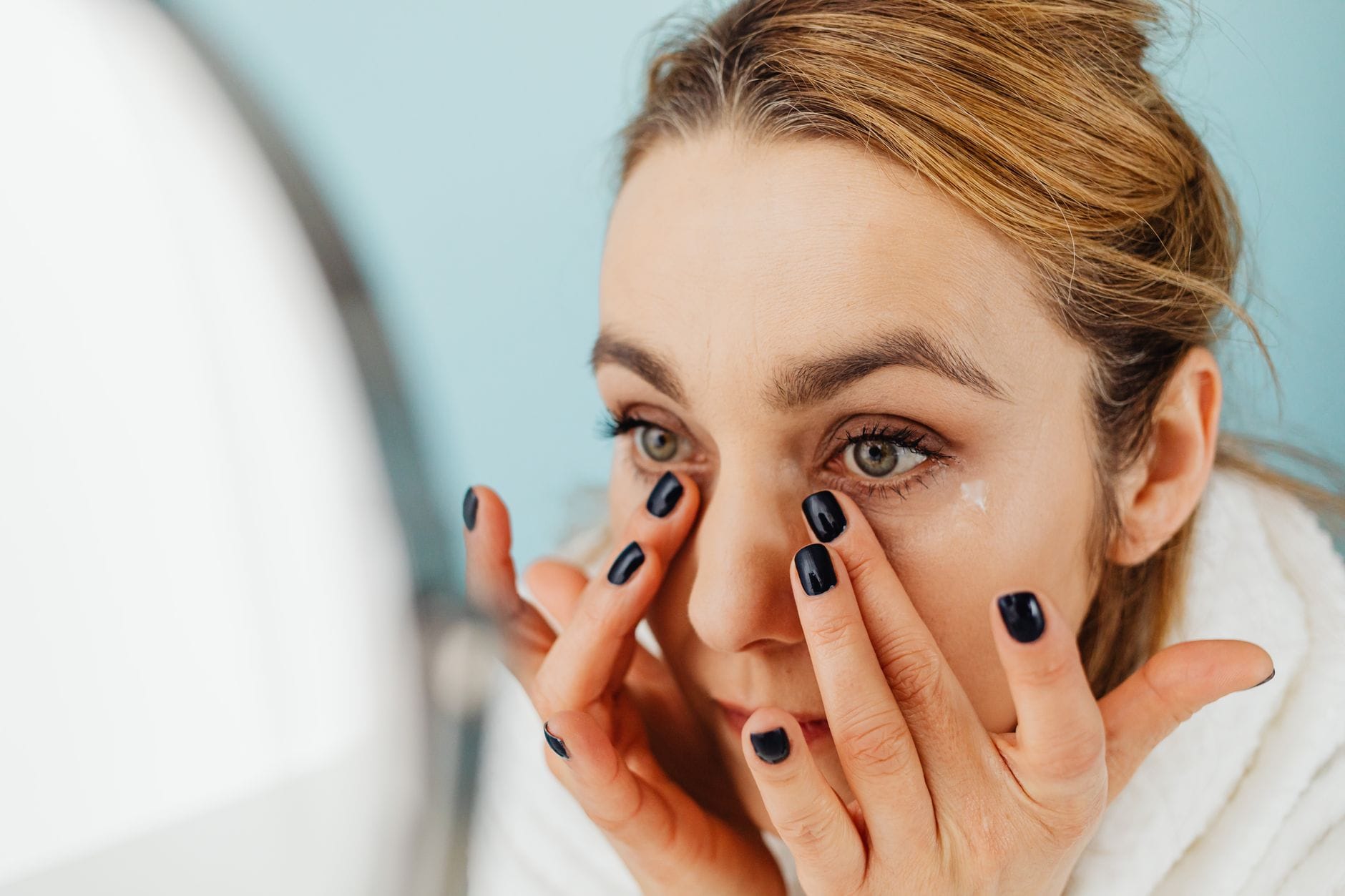 Pielęgnacja skóry wokół oczu: co ma sens, a co jest marketingiem Kobieta nakłada krem pod oczy, patrząc w lustro na niebieskim tle